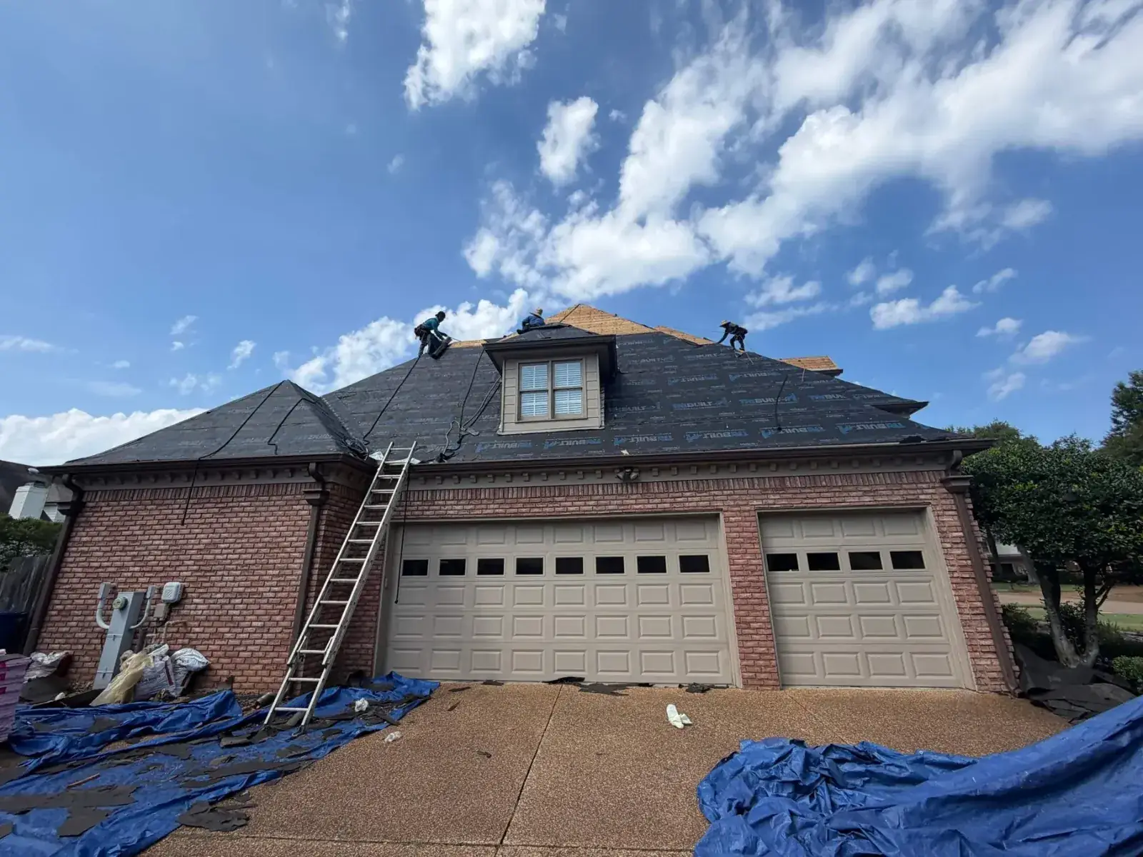 Crew installing synthetic underlayment on a brick home over a three-car garage, dramatic blue sky and clouds