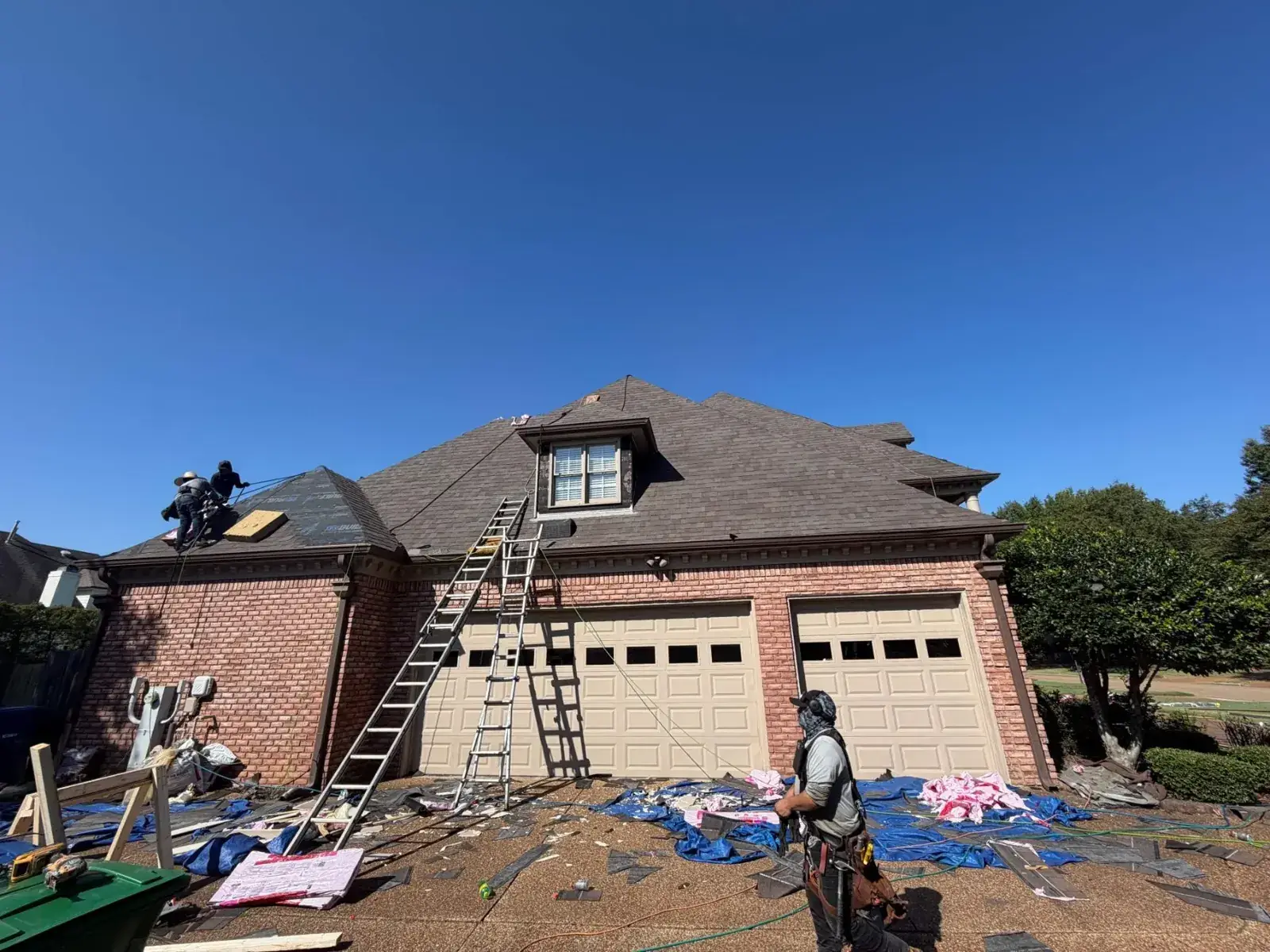 Roofers on a brick home's roof during tear-off while a harnessed crew member (face covered) works the tarped driveway