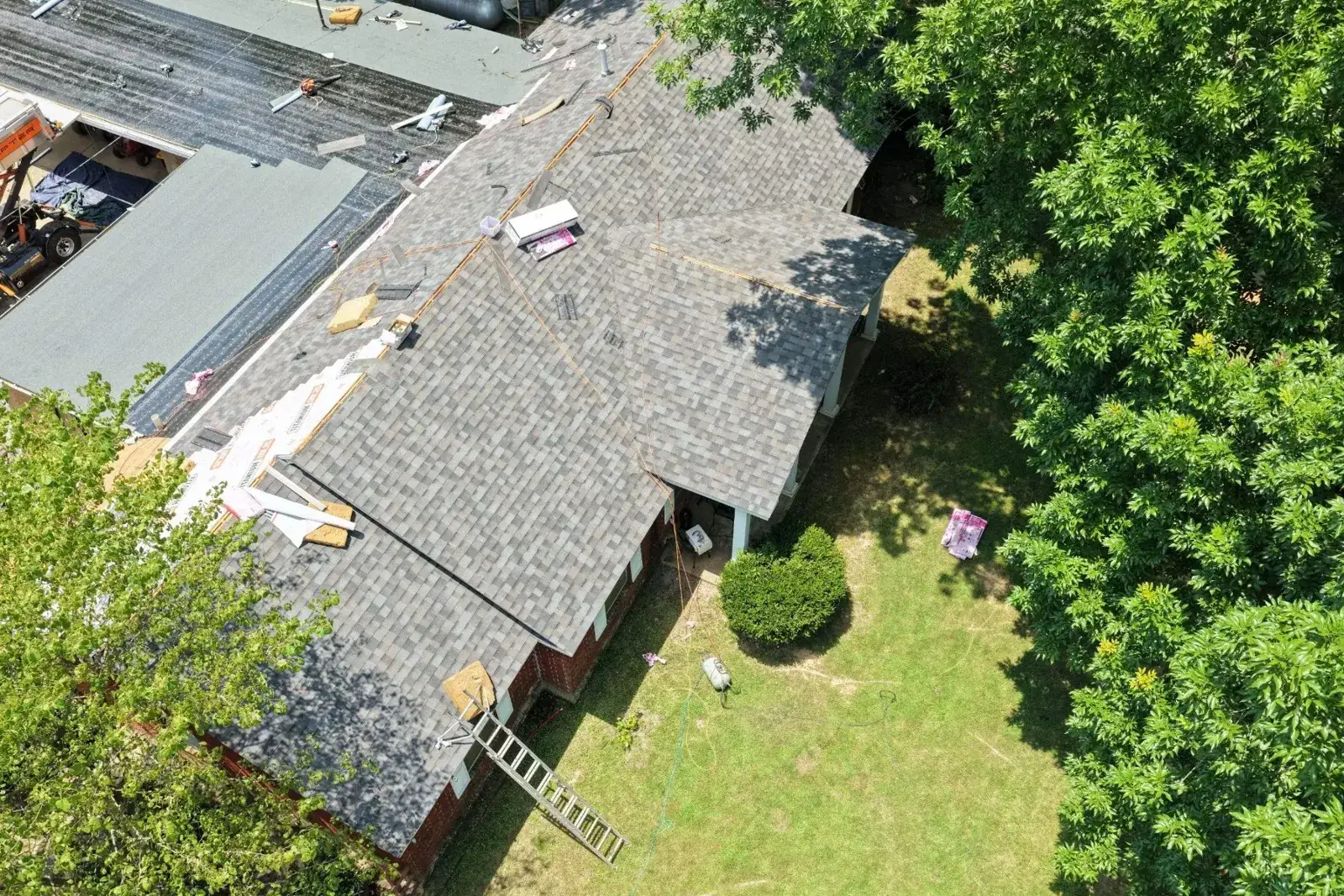 Drone overhead of a brick ranch mid-install: new shingles on one wing, flat-roof section and crew materials staged