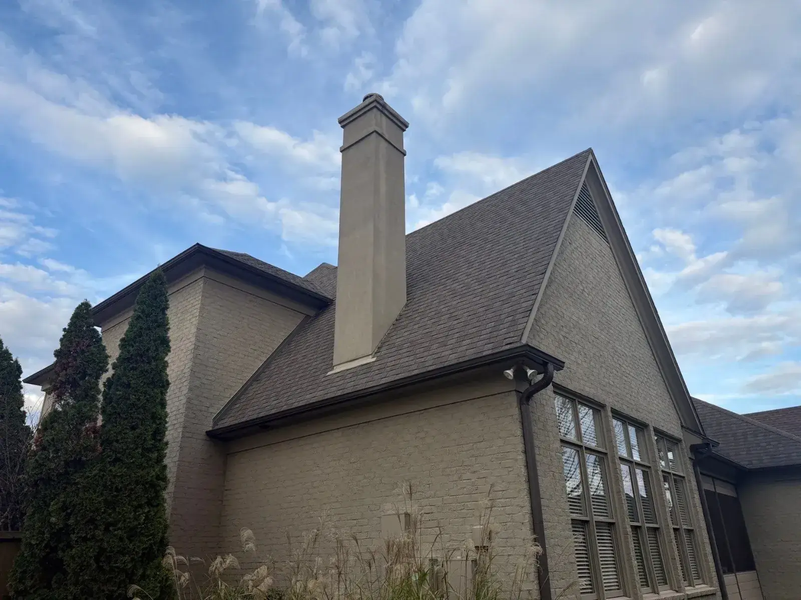 Upscale painted-brick home with steep new shingle roof, tall stucco chimney and gable vent against a soft blue sky