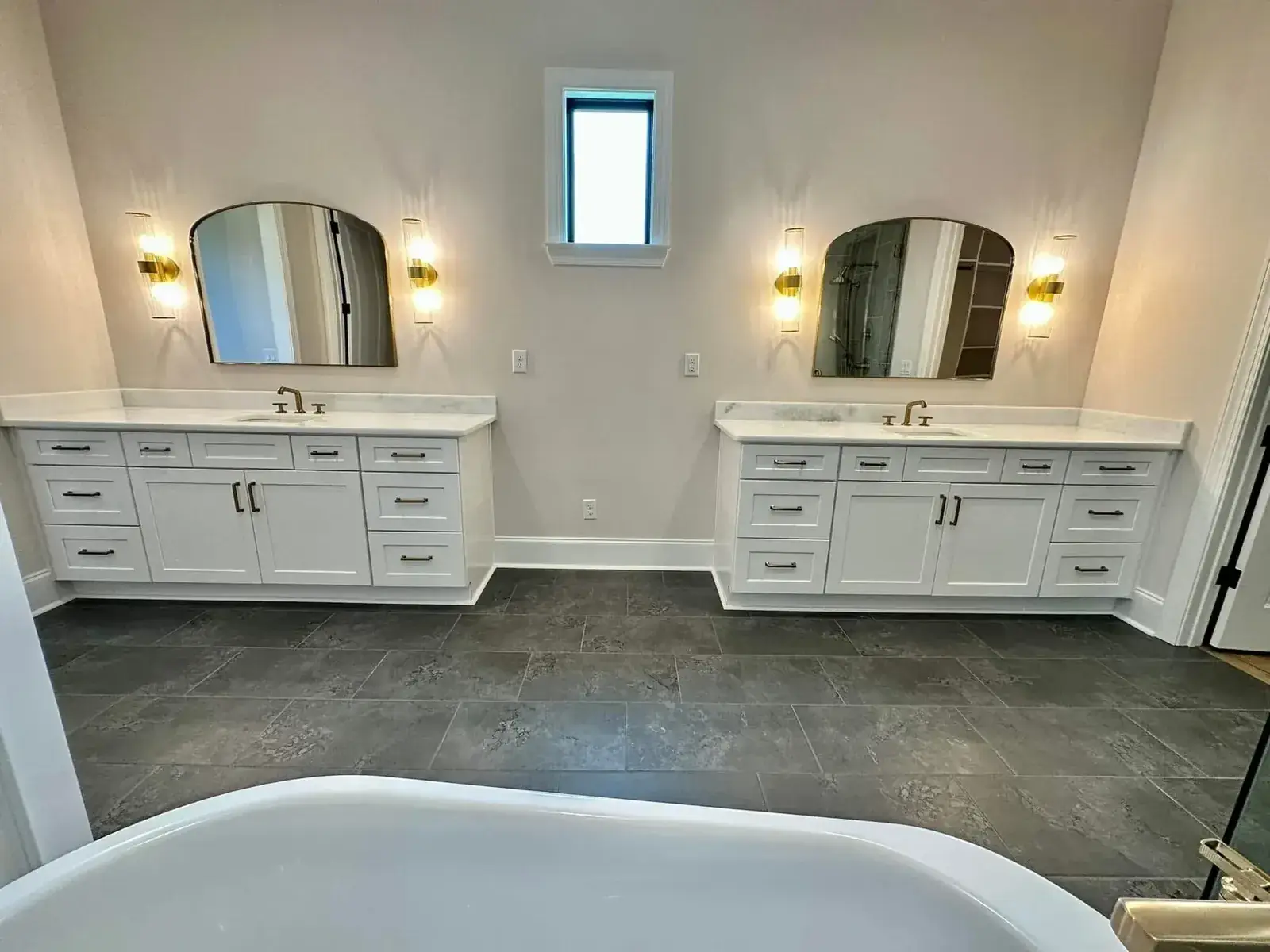 Twin white vanities with arched mirrors and brass sconces viewed from a freestanding tub, dark tile floor