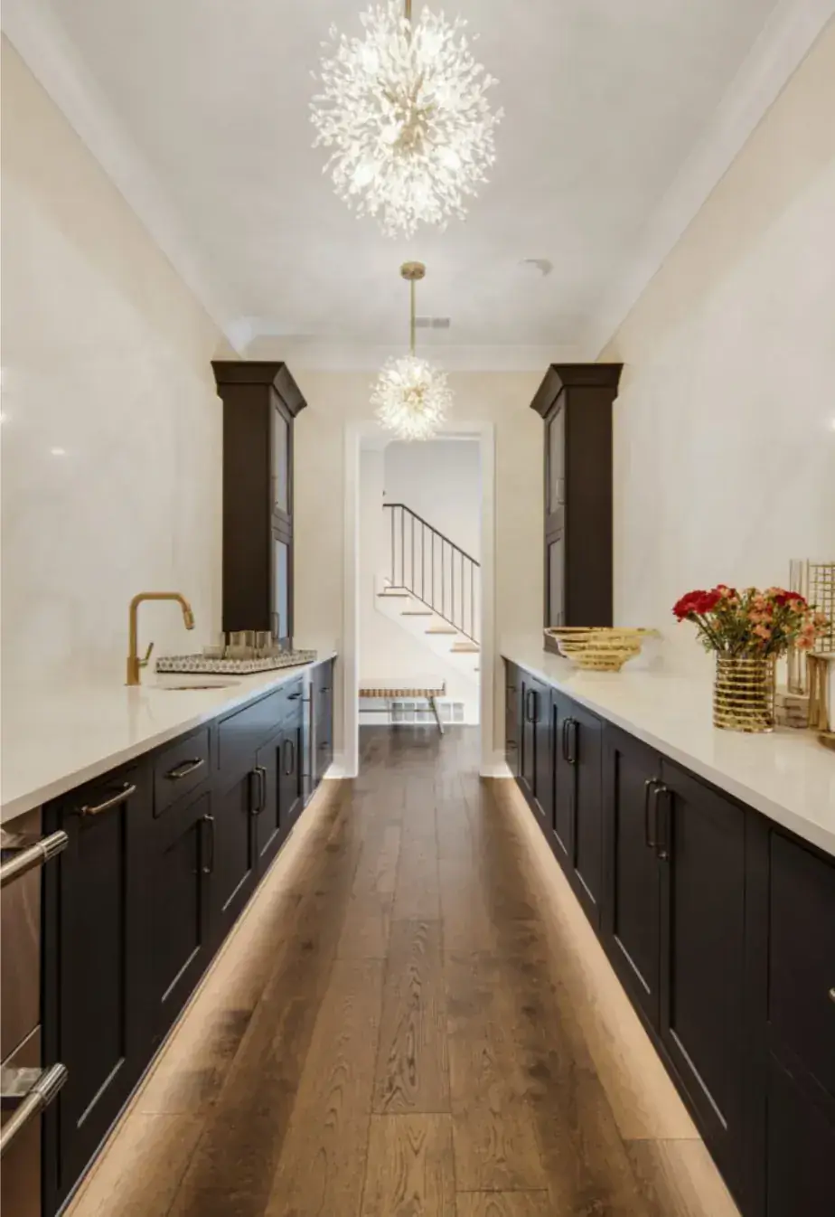 Galley butler's pantry with dark cabinetry, slab-marble walls, brass faucet, sparkle chandeliers and lit wood floors
