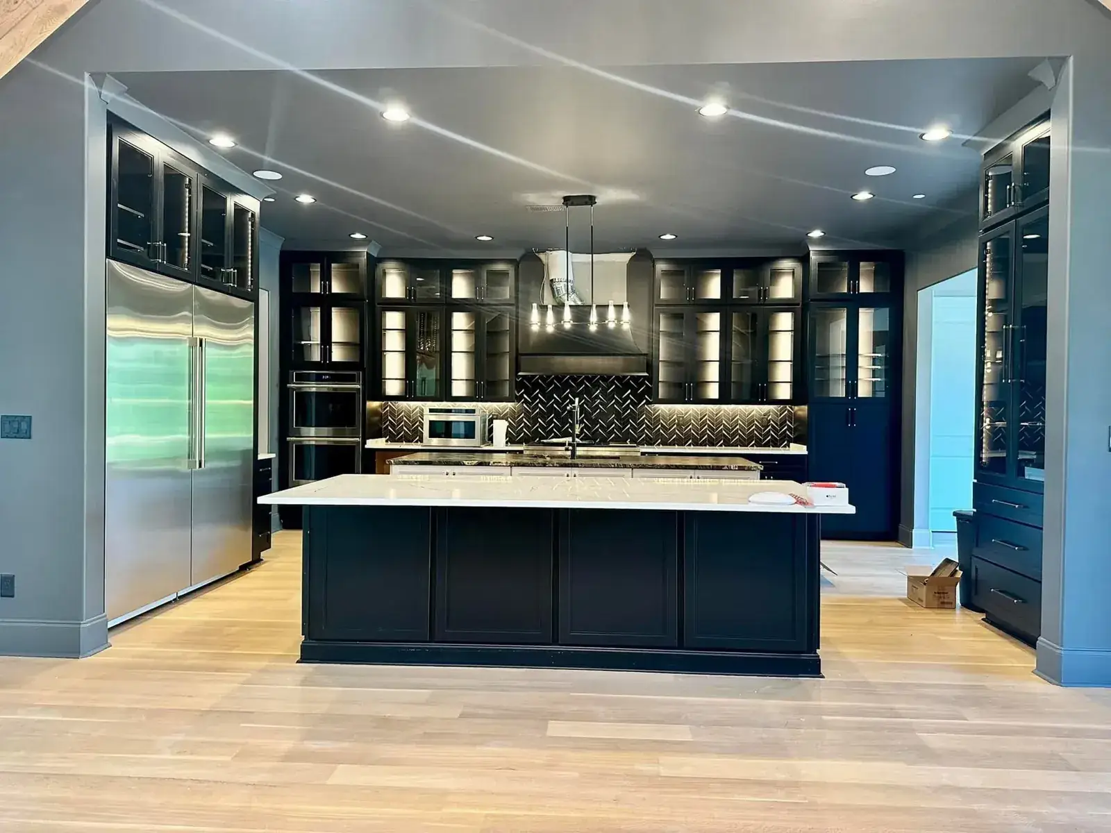 Dramatic kitchen with black glass-front lit cabinetry, chevron backsplash, large island and built-in refrigerator