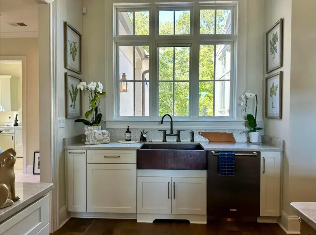 Hammered copper farm sink beneath a wall of windows, white shaker cabinets, orchids and botanical prints