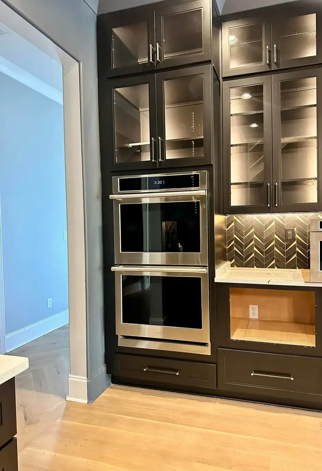Double wall ovens framed by dark lit glass-front cabinets and chevron tile coffee nook