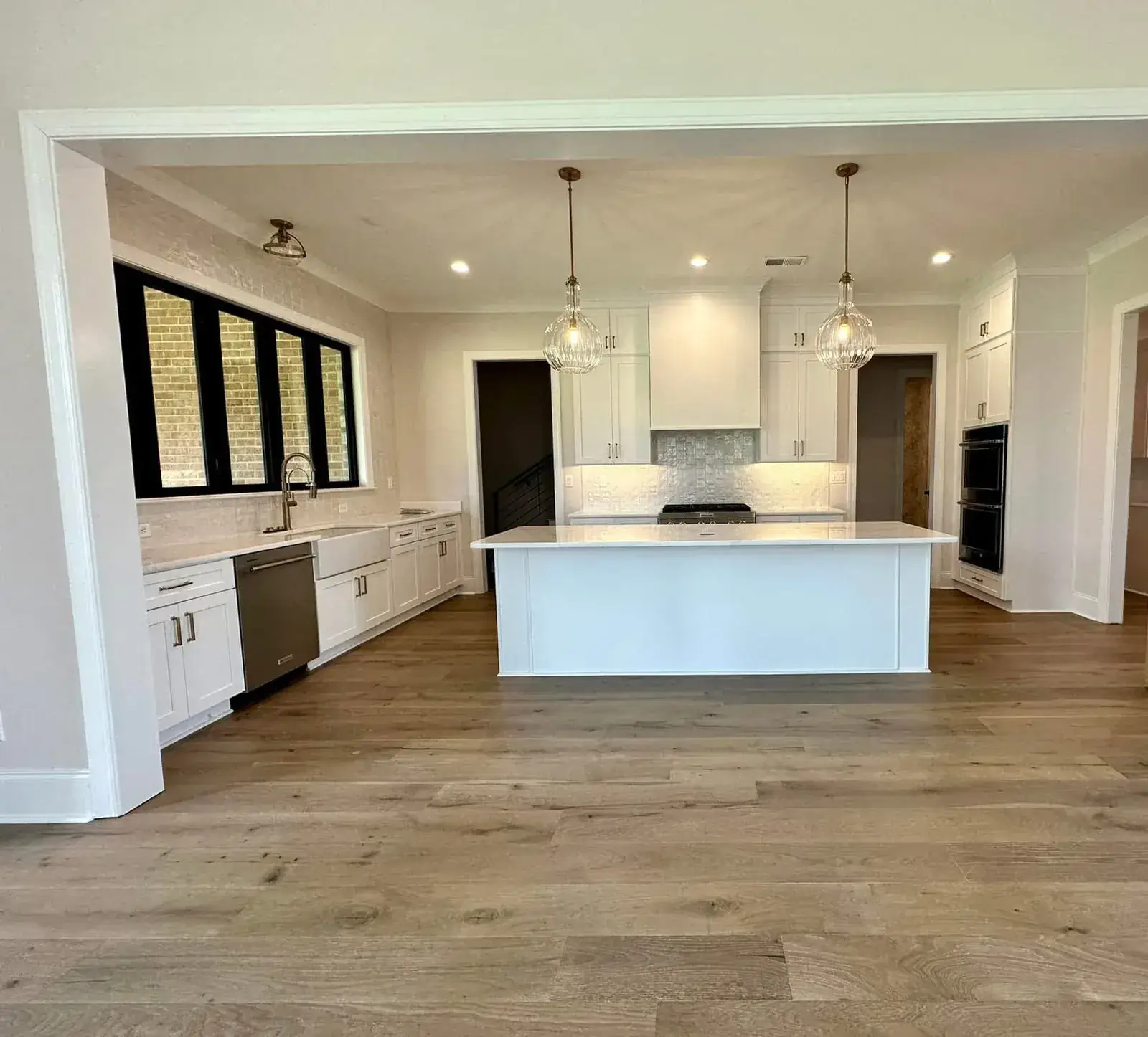 Open-plan white kitchen with island, ribbed-glass pendants, farm sink under black windows and wide white-oak floors