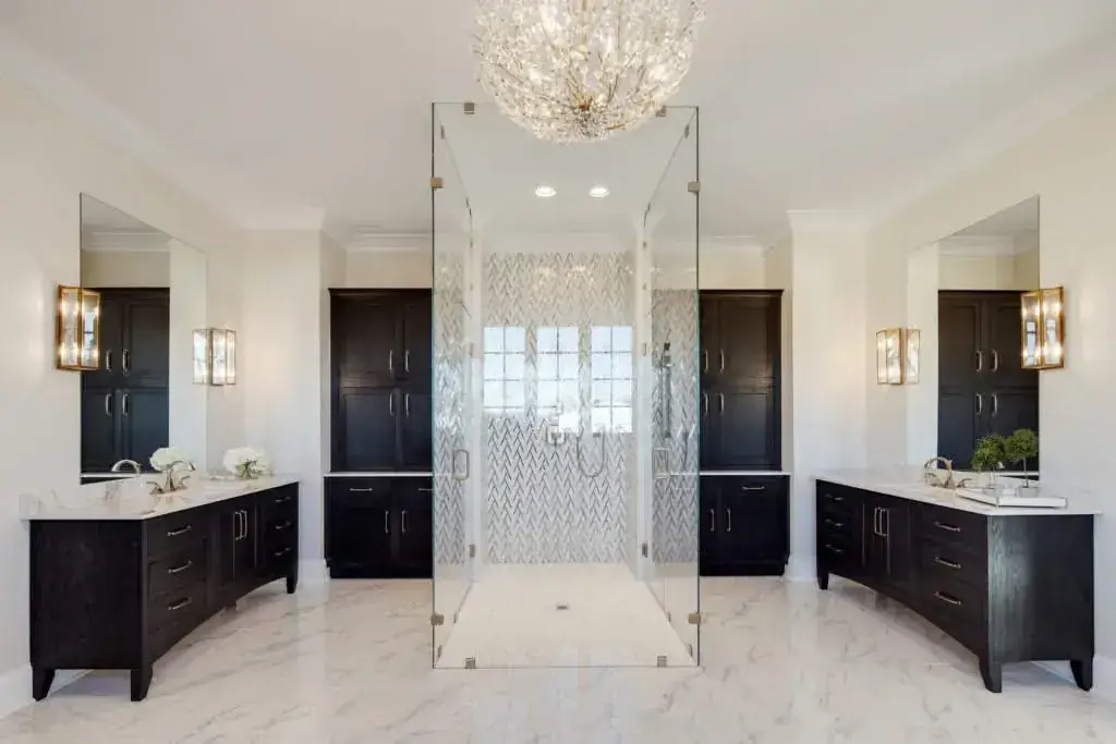 Symmetrical luxury bath with walk-through frameless glass shower, herringbone tile, dark vanities and crystal chandelier
