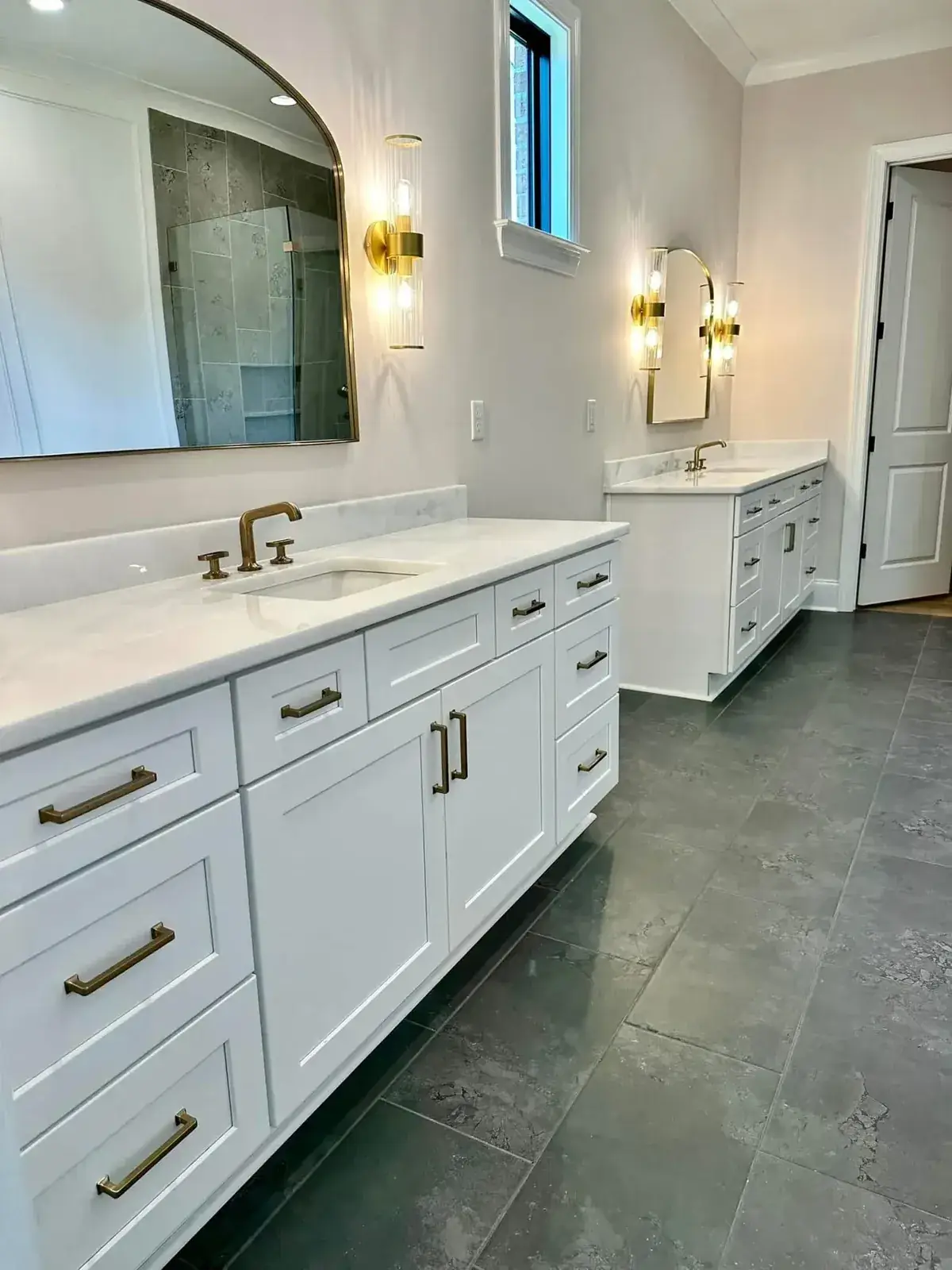 Primary bath with white shaker double vanities, marble tops, brushed-brass faucets and sconces, and slate-gray tile floor