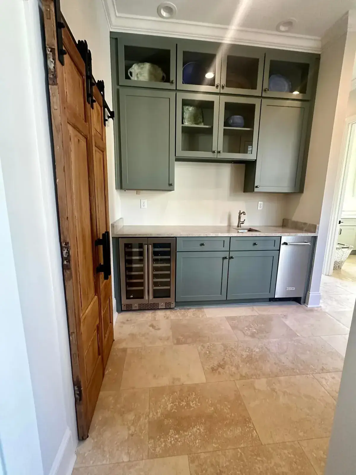 Sage wet bar with glass-front uppers, wine fridge, travertine floor and reclaimed-wood barn door